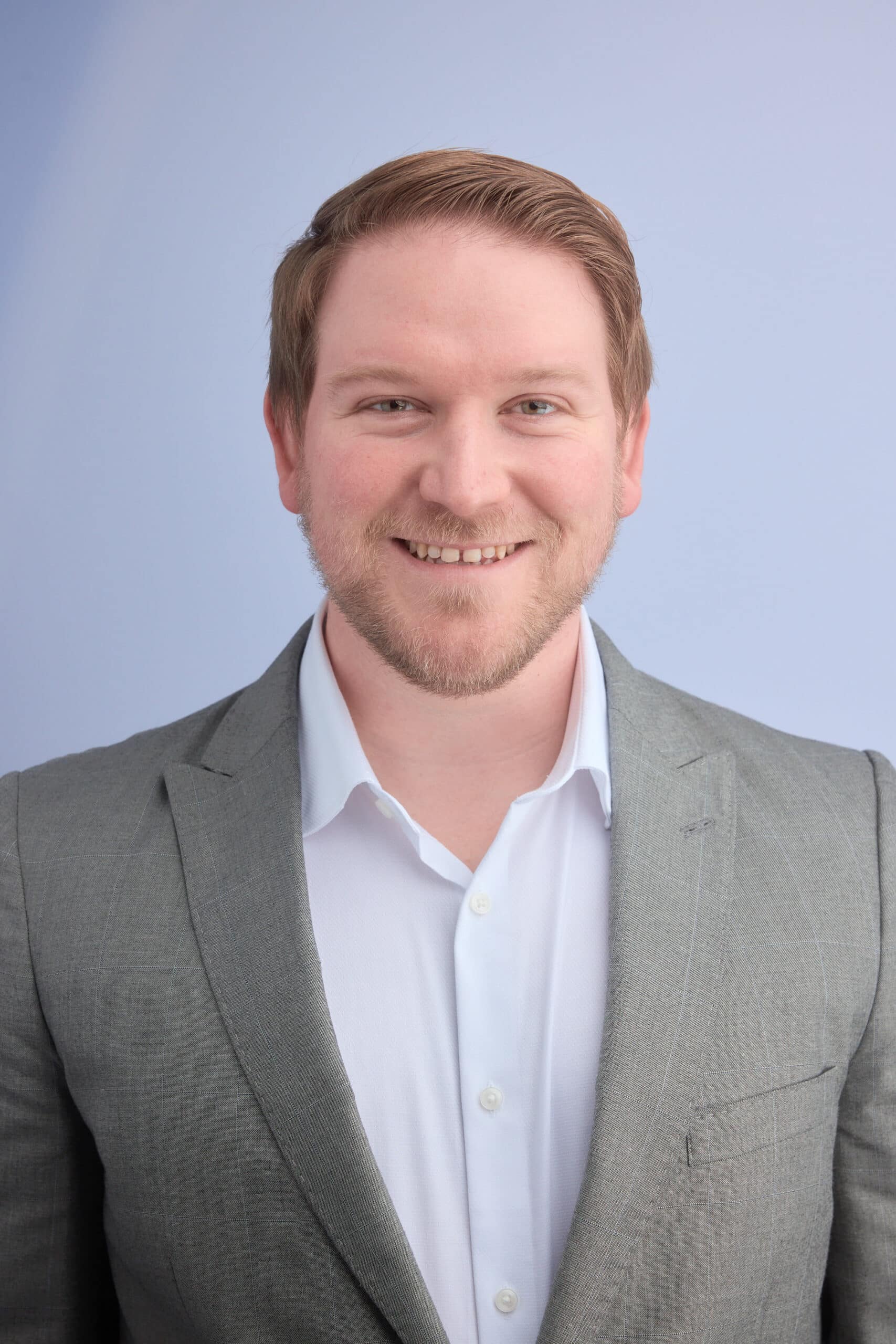 Upper-body portrait of a smiling man in a light gray suit and white shirt against a blue background.