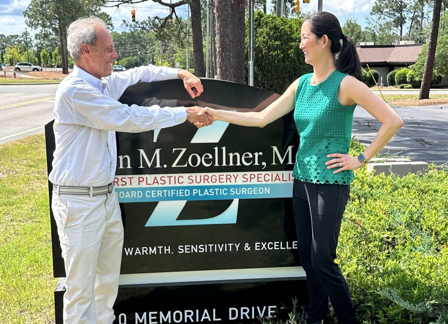 Two adults shake hands in front of a plastic surgery clinic sign outdoors on a sunny day, smiling at each other.