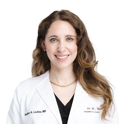 Smiling female physician in a white lab coat over a black top, wearing a gold chain, against a white background.