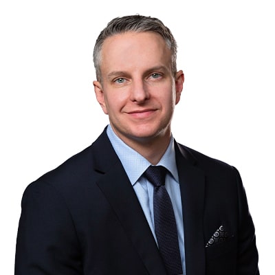 Professional headshot of a smiling man in a navy suit, light blue shirt, and dark tie, against a white background.