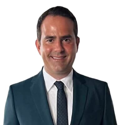 Head-and-shoulders portrait of a smiling man in a dark teal suit, white shirt, and navy tie against a white background.