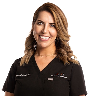 Smiling female cosmetic surgeon in black scrubs with HKB Cosmetic Surgery logo on chest.