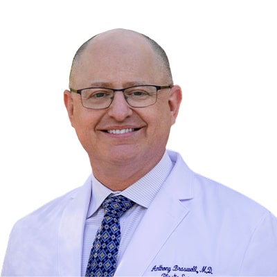 Smiling male doctor in a white lab coat and patterned blue tie, wearing glasses, posing for a portrait.
