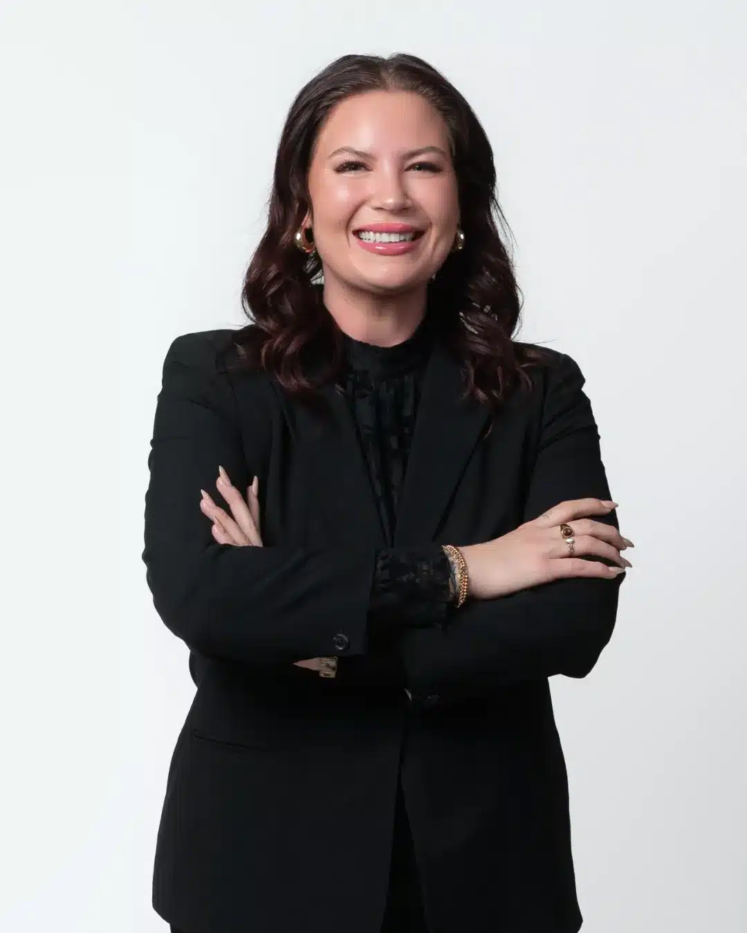 Professional woman smiling with arms crossed, wearing a black blazer and gold jewelry against a light background.