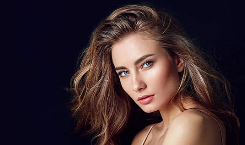 Portrait of a woman with wavy light brown hair and subtle makeup, posed against a dark background, looking at the camera.