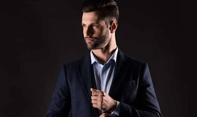 Man in a dark suit adjusts his cuff while looking to the left in a studio with a dark background.