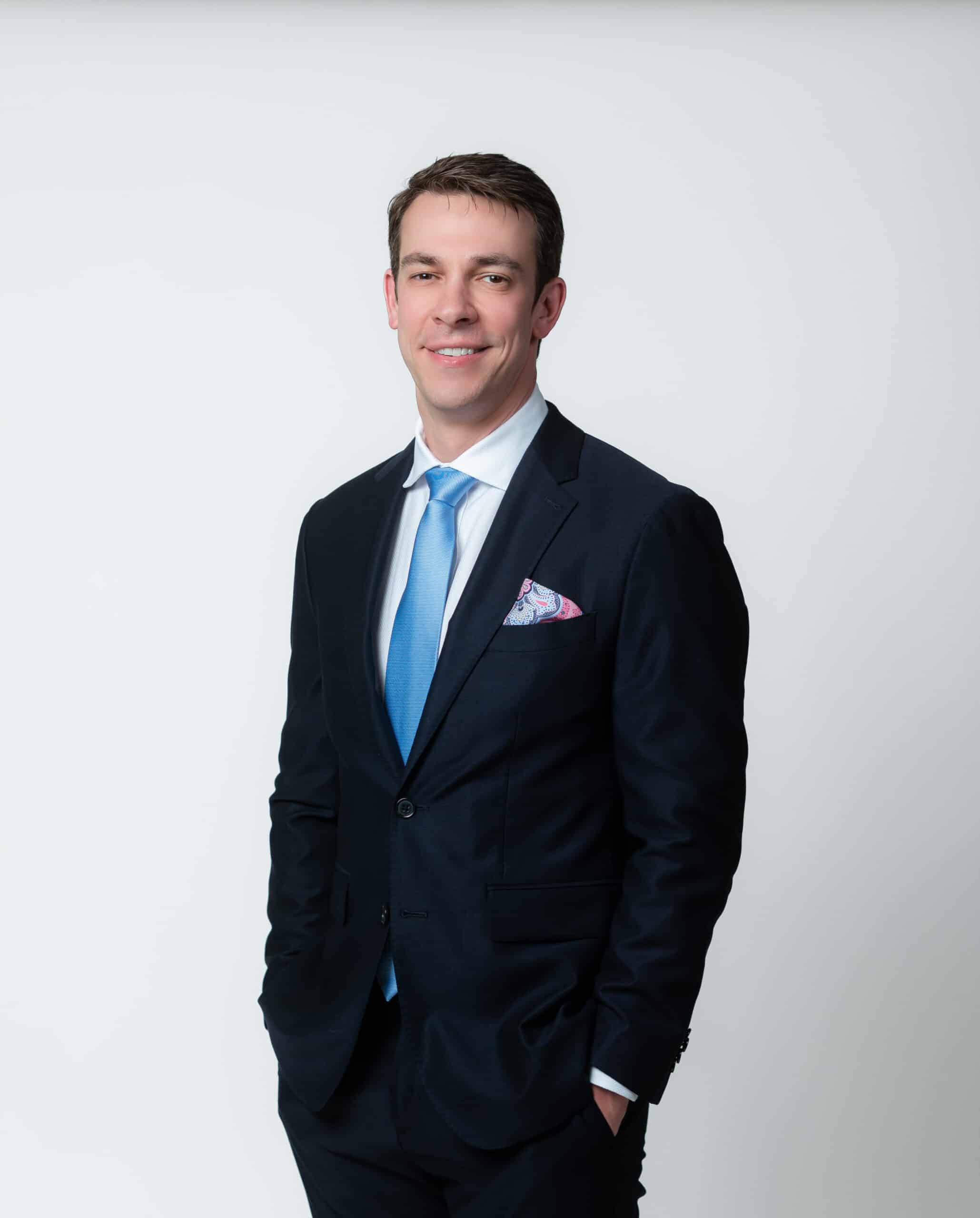 Professional man in a dark navy suit with a light blue tie, smiling against a light gray studio background, hands in pockets