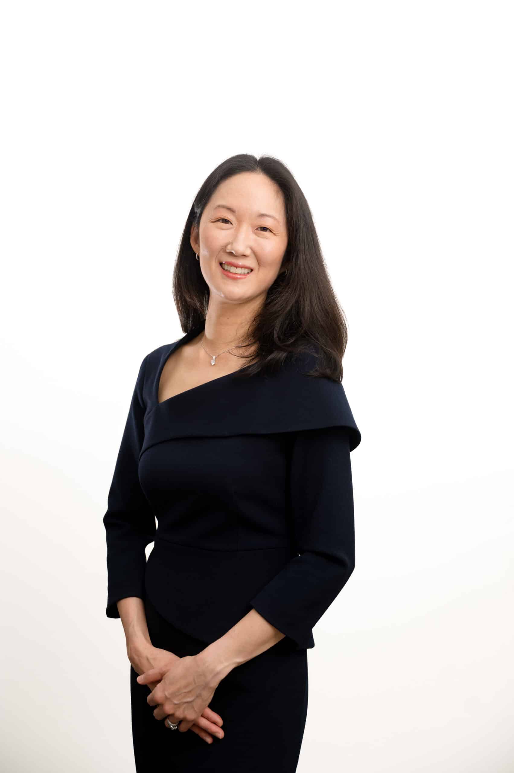 Professional portrait of a smiling woman in a navy dress with hands clasped in front, white studio background.