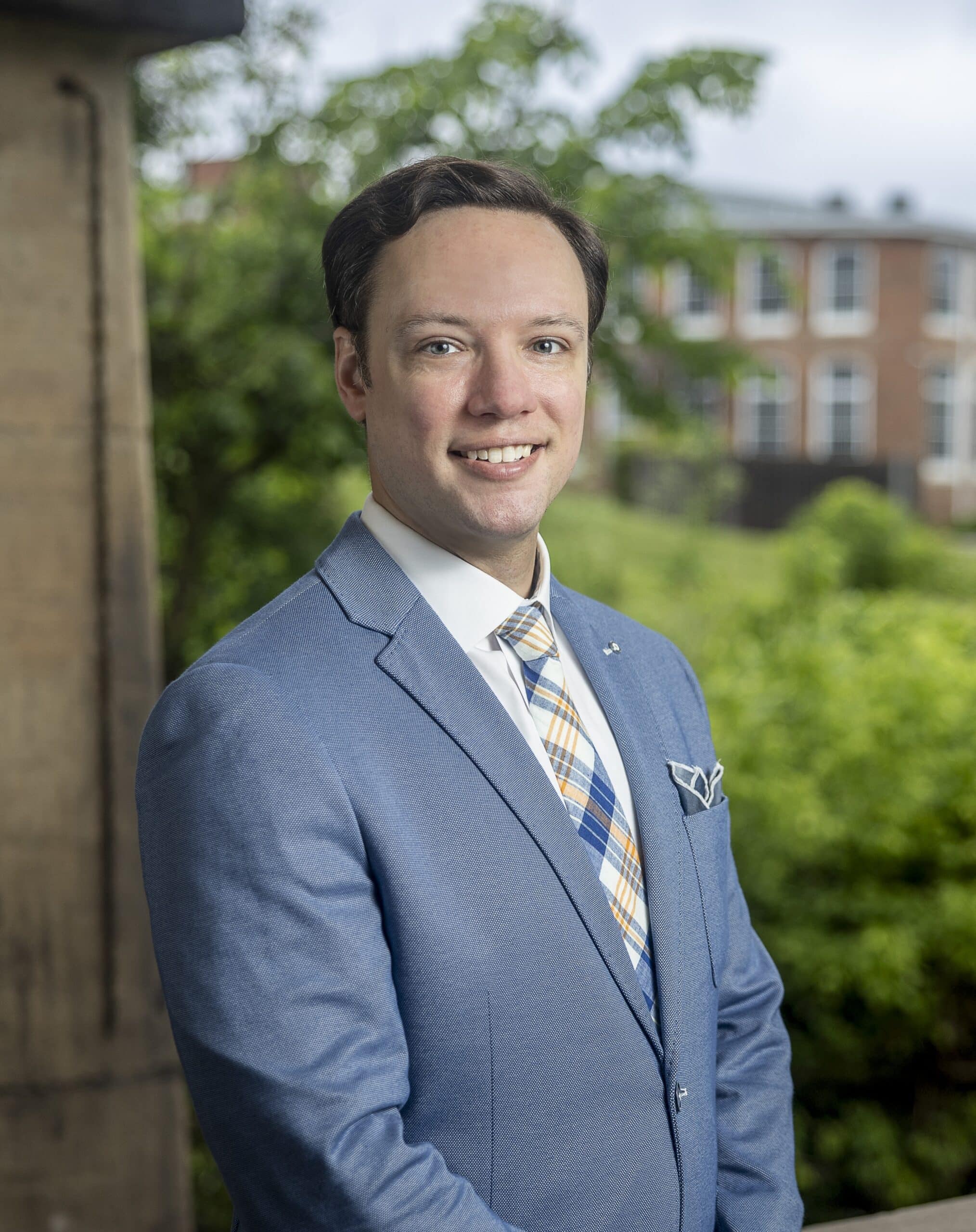 Portrait of a smiling man in a light blue suit with a plaid tie, standing outdoors with greenery and buildings in the background.