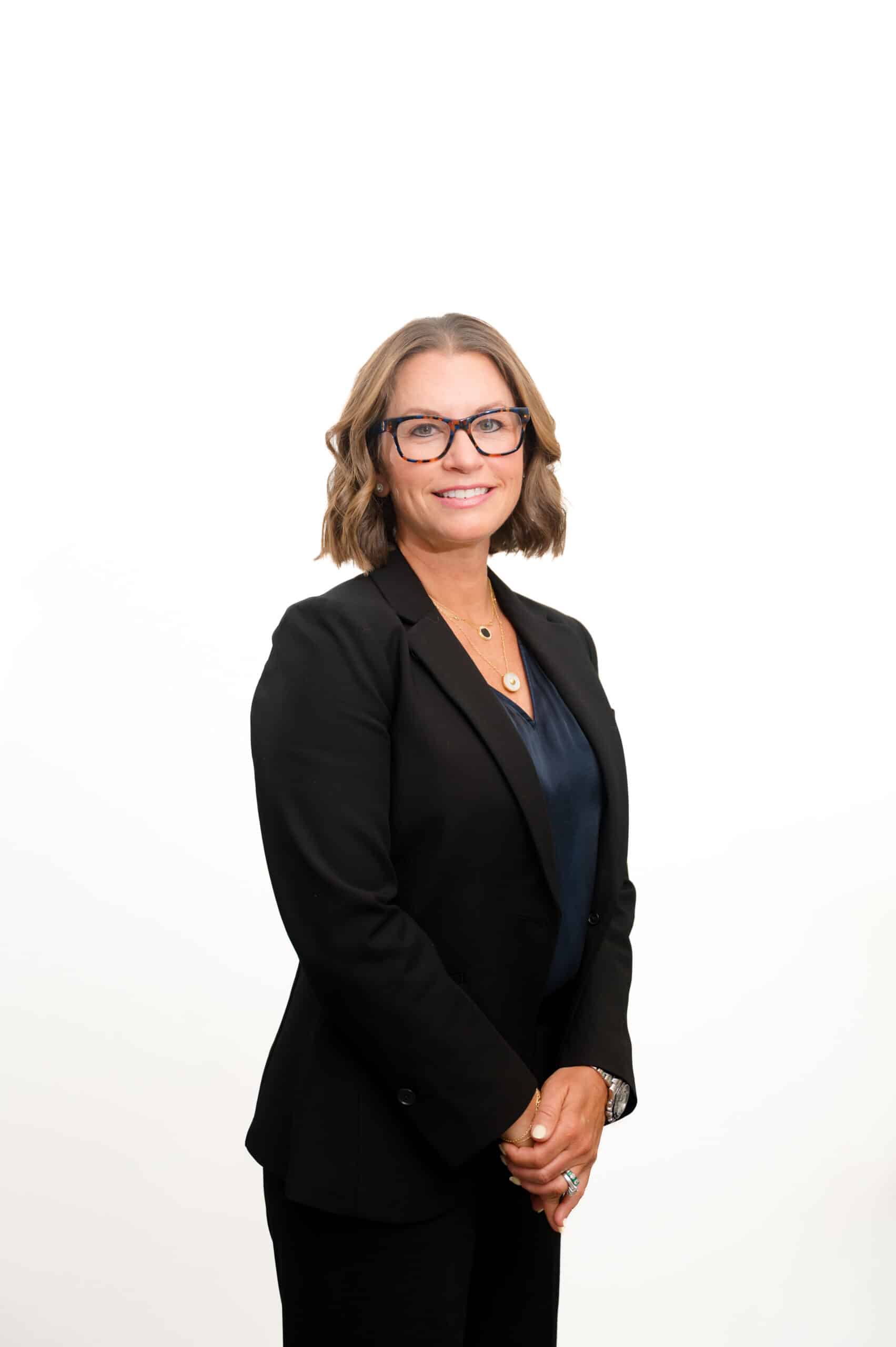 Professional woman in a black blazer and navy blouse, wearing tortoiseshell glasses, smiling.