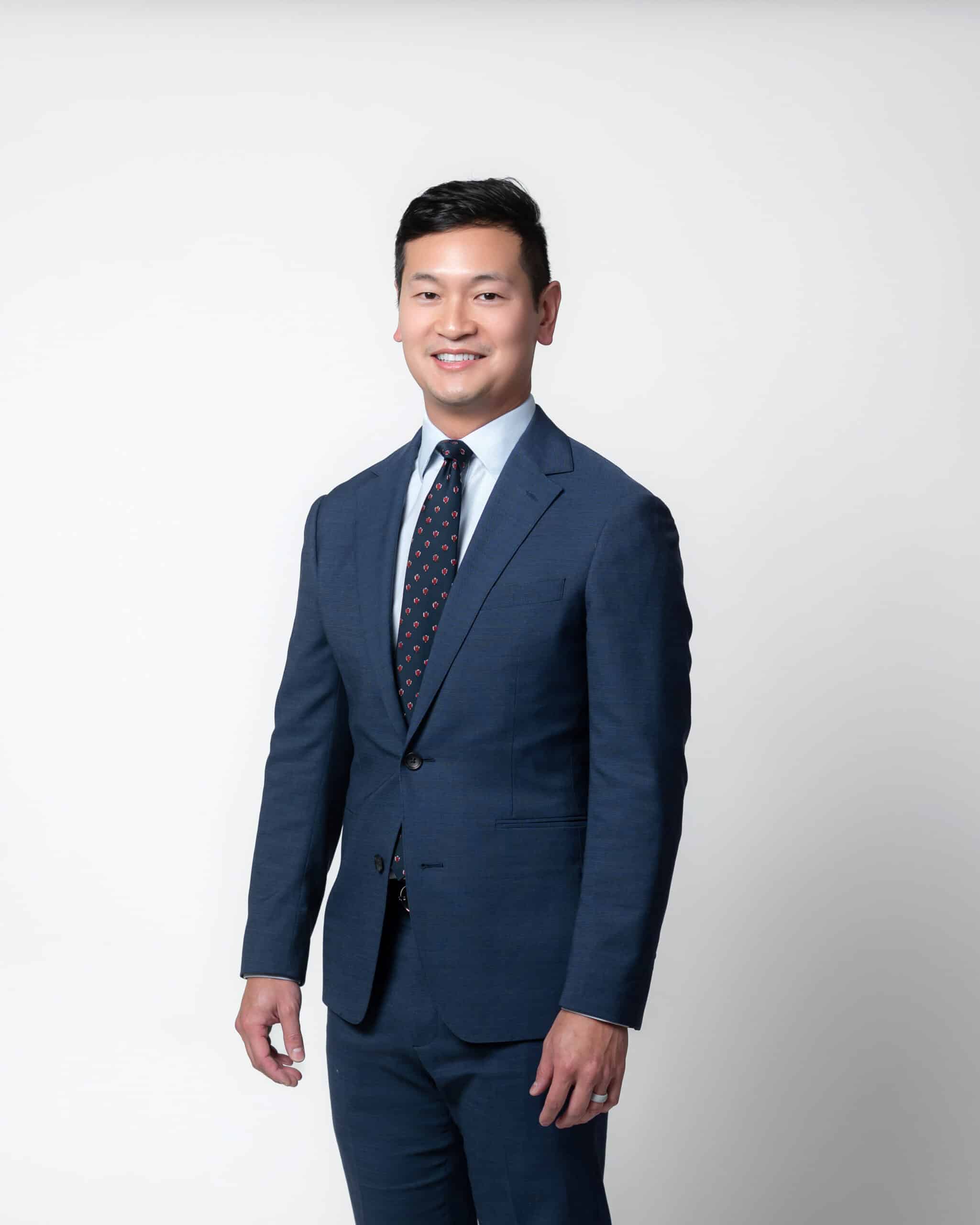 Portrait of a smiling man in a navy suit and patterned tie against a light background.