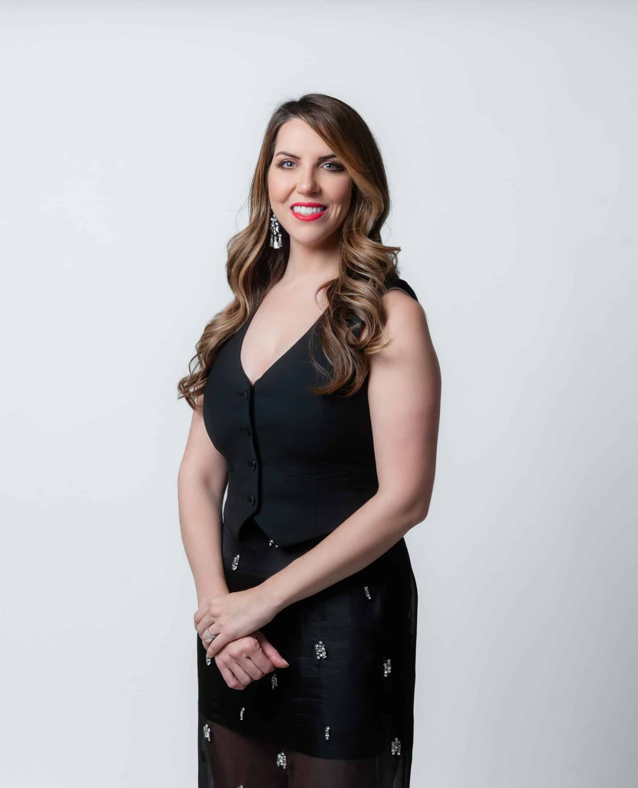 Smiling woman in a black sleeveless dress with rhinestone details, standing against a light gray backdrop.Long, wavy brown hair frames her face as she looks at the camera.