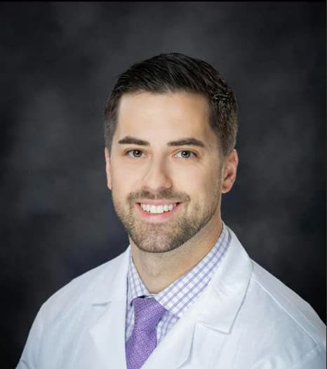 Portrait of a smiling man in a white coat and purple tie against a dark backdrop, portraying a medical professional.