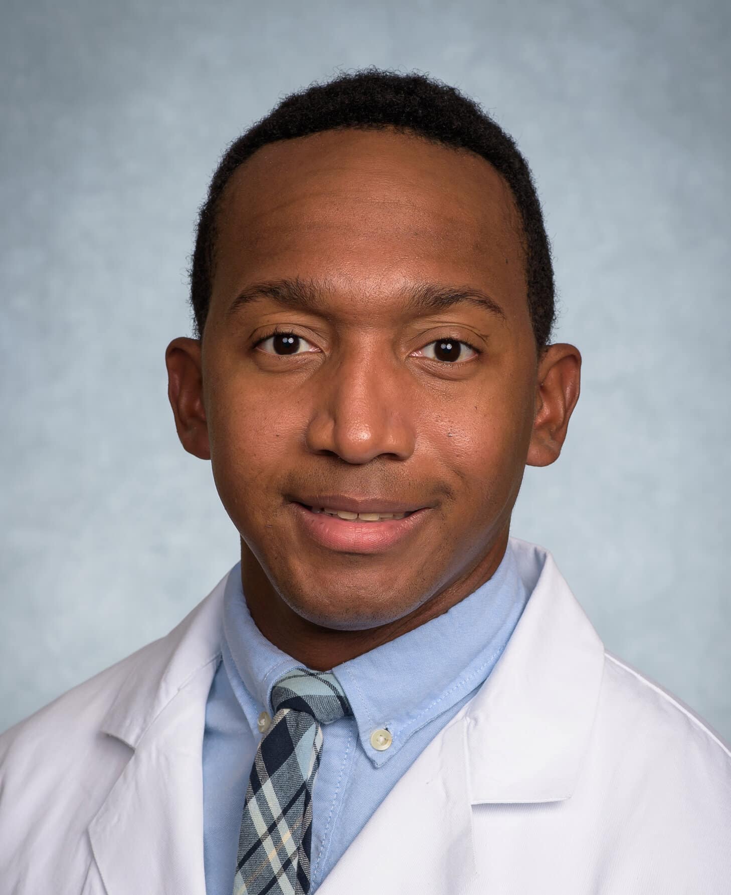 Portrait of a man in a white lab coat and blue shirt with a plaid tie, smiling softly against a pale blue background.