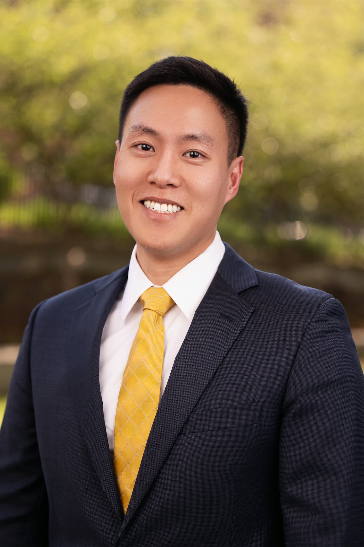 Portrait of a smiling man in a navy suit with a yellow tie, outdoors with a green, blurred background.
