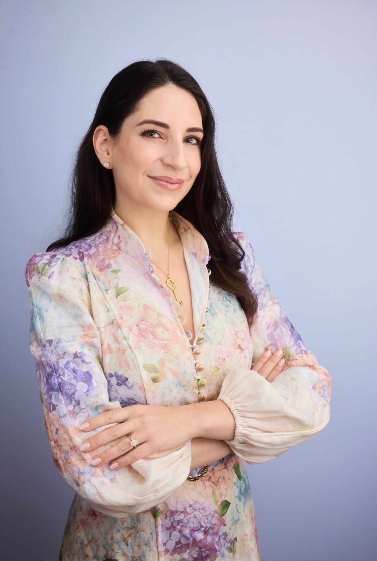 Professional-looking woman with long dark hair standing against a light blue background, wearing a pastel floral blouse and a gold pendant, arms crossed.