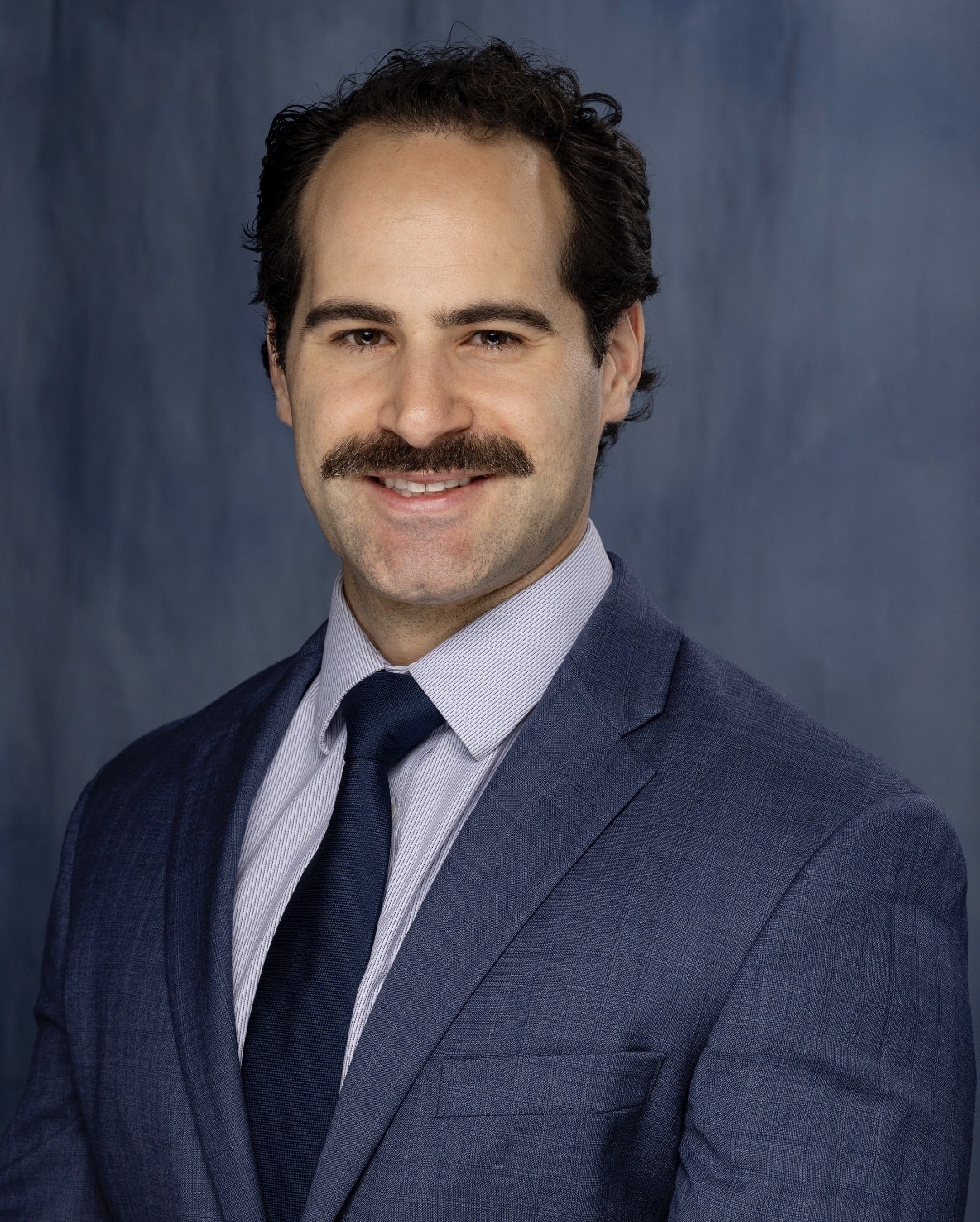 Smiling man in a blue suit and tie posing for a formal portrait against a blue backdrop.